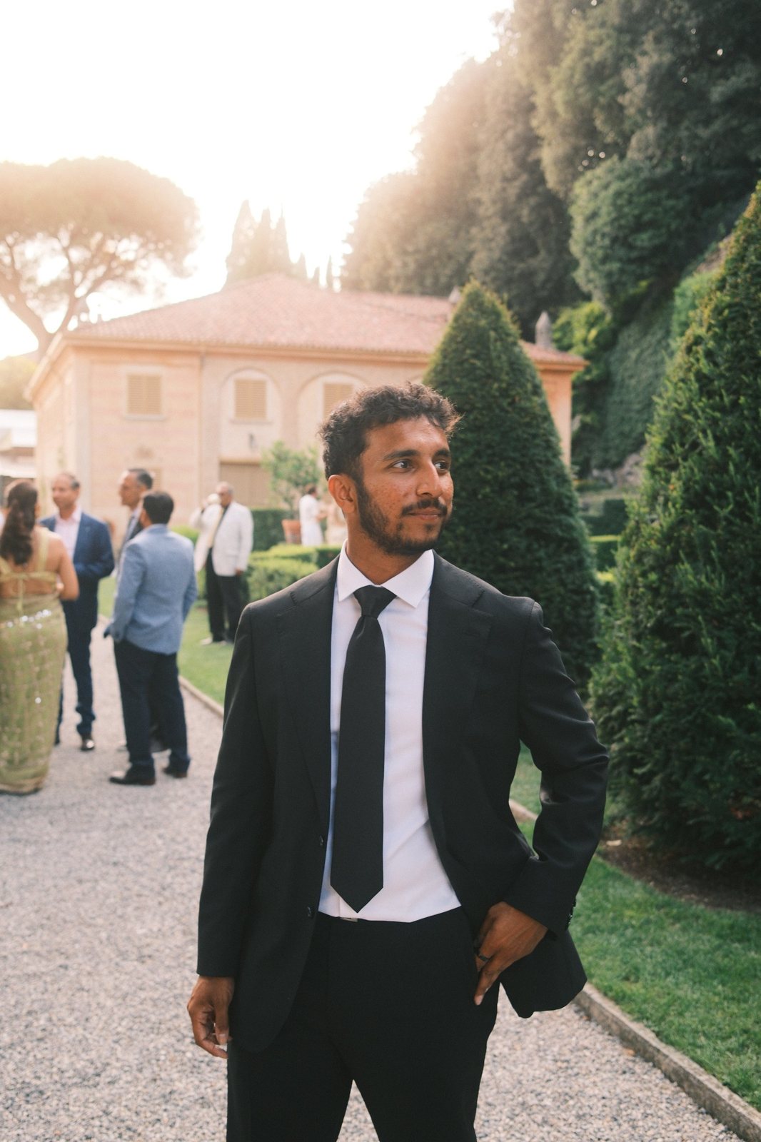 Manav Parekh in a black suit in a sunlit Italian garden, smiling and looking off to the side