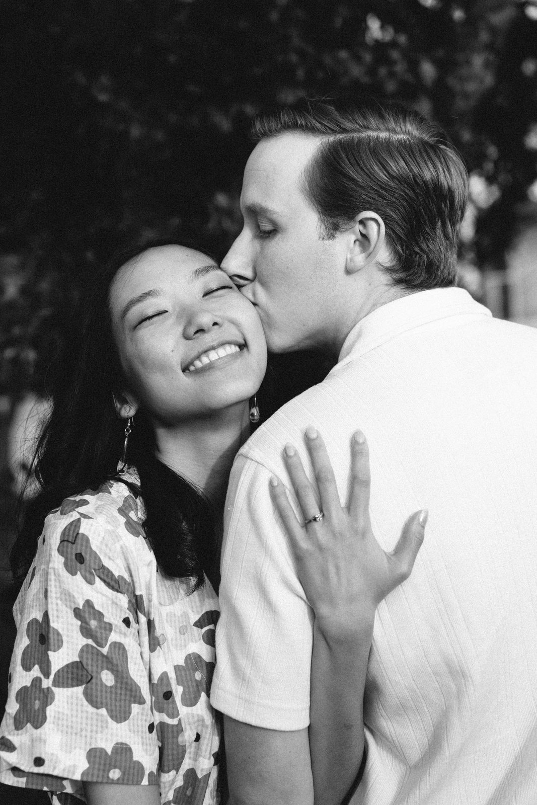 Black and white photo of him kissing her cheek as she smiles, ring hand on his shoulder