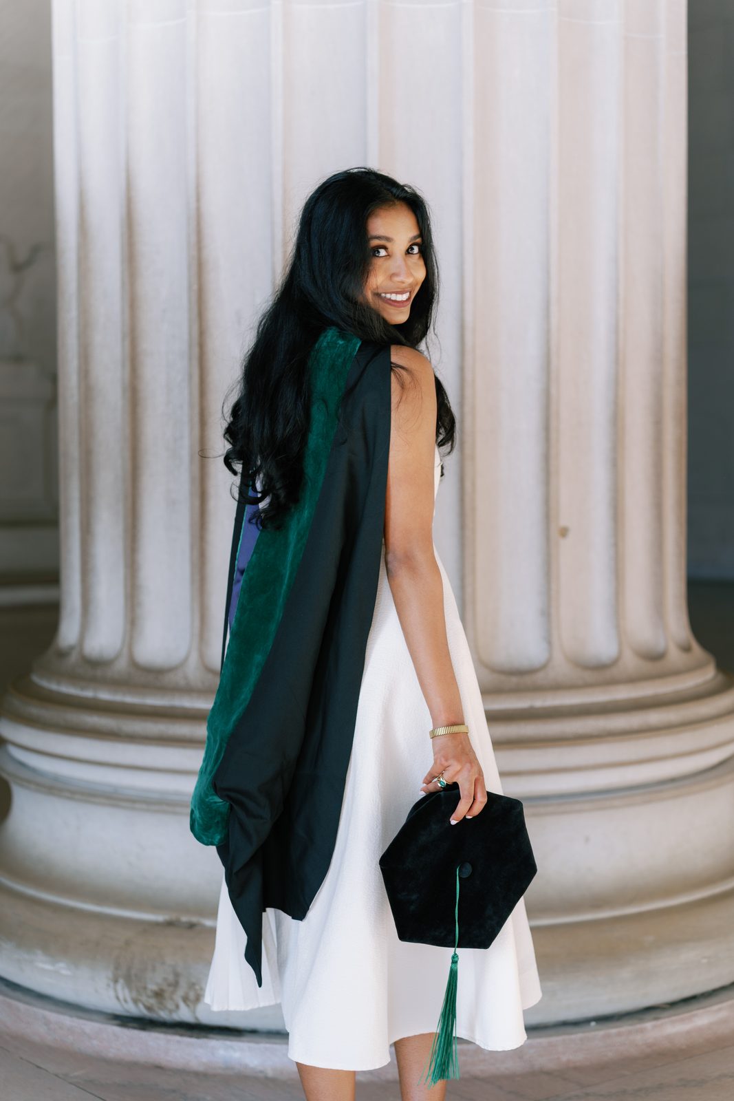 Graduate in a green hood looking back over her shoulder beside a white column, cap in hand