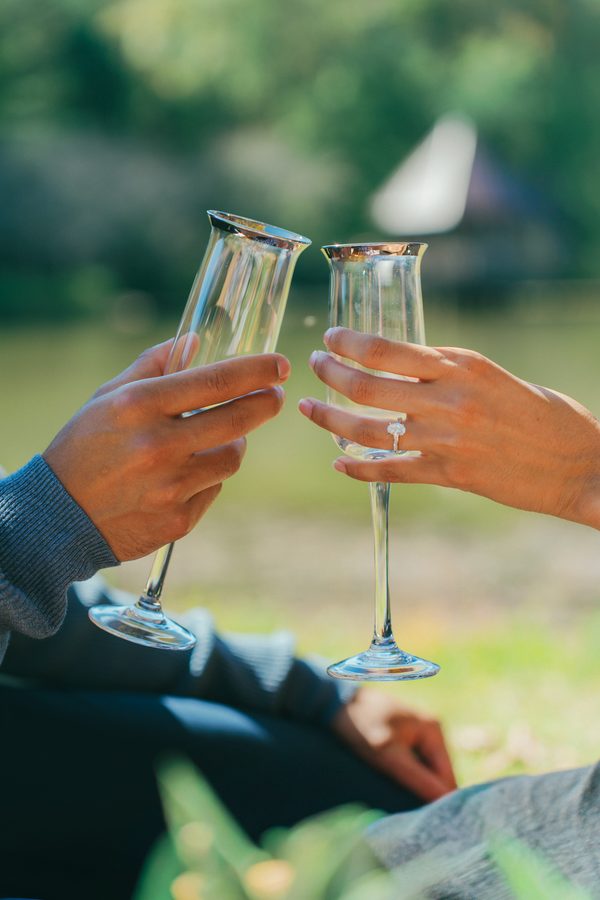 Two champagne flutes clinking by a lake, engagement ring in view