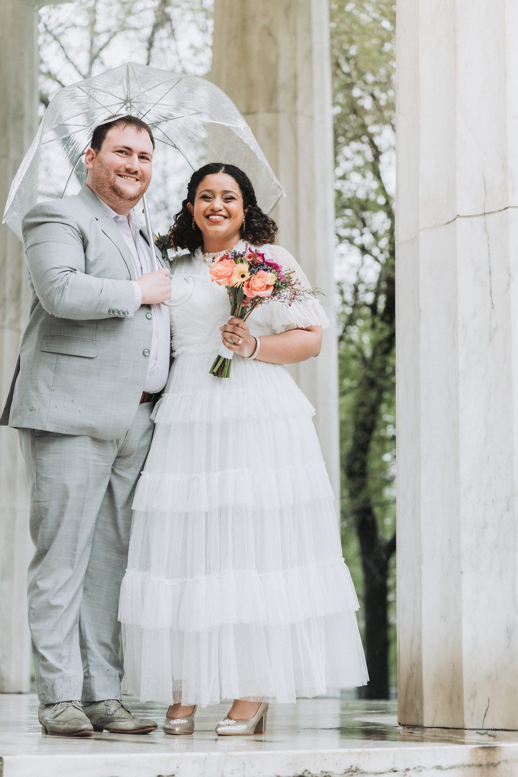 Bride and groom under a clear umbrella between white marble columns on a rainy day